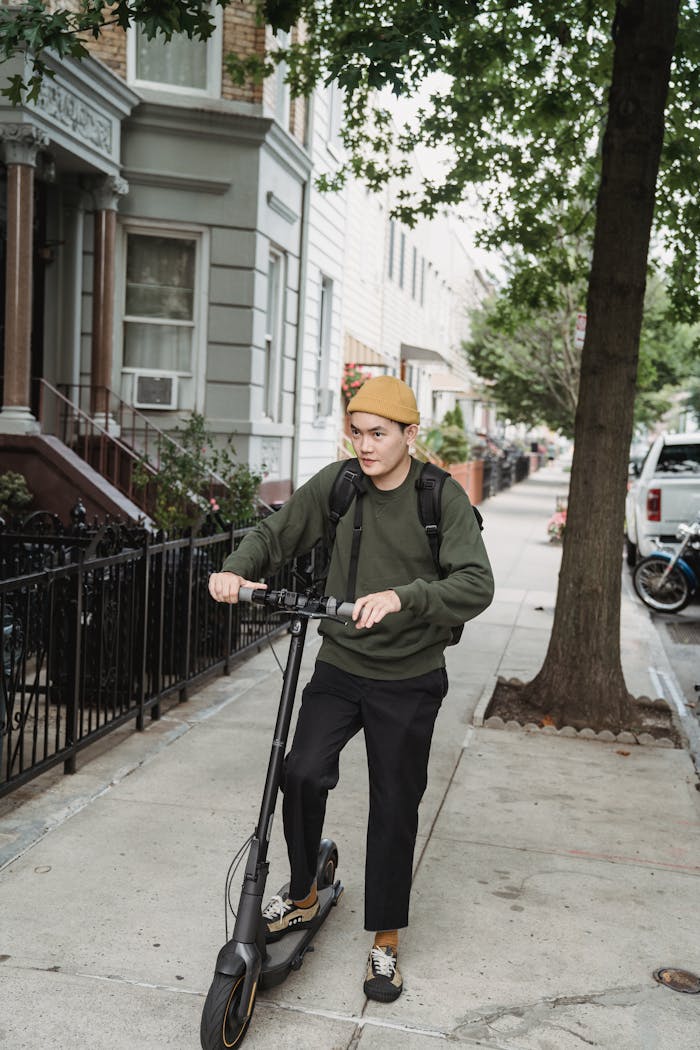 A young man in casual attire rides an electric scooter on a tree-lined sidewalk.