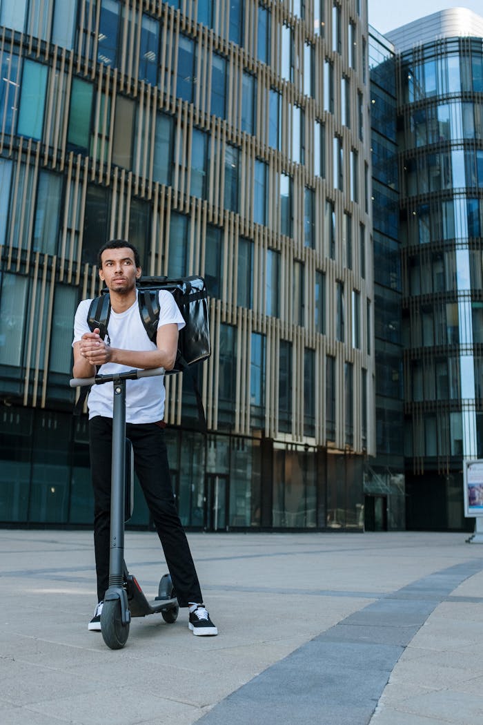 Young man with backpack and e-scooter in front of a modern office building, representing sustainable urban delivery.